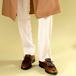 Cognac brown shoes with fringe by Timothée Paris worn with creme pants and a brown coat on a beige background