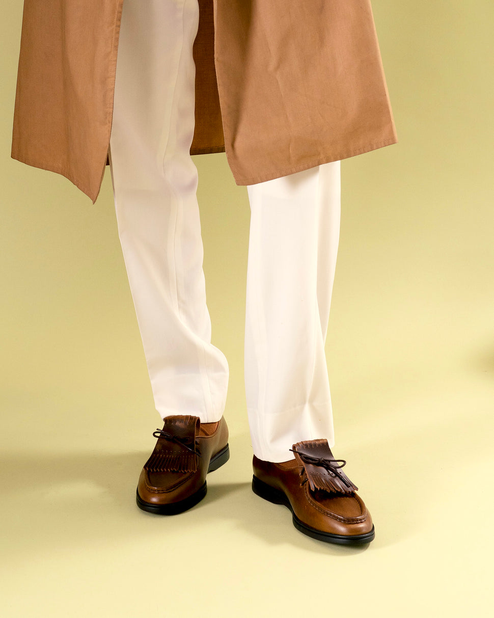 Cognac brown shoes with fringe by Timothée Paris worn with creme pants and a brown coat on a beige background