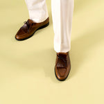 Relaxed brown loafers with tassels worn with white pants on a pistachio green background Timothée Paris