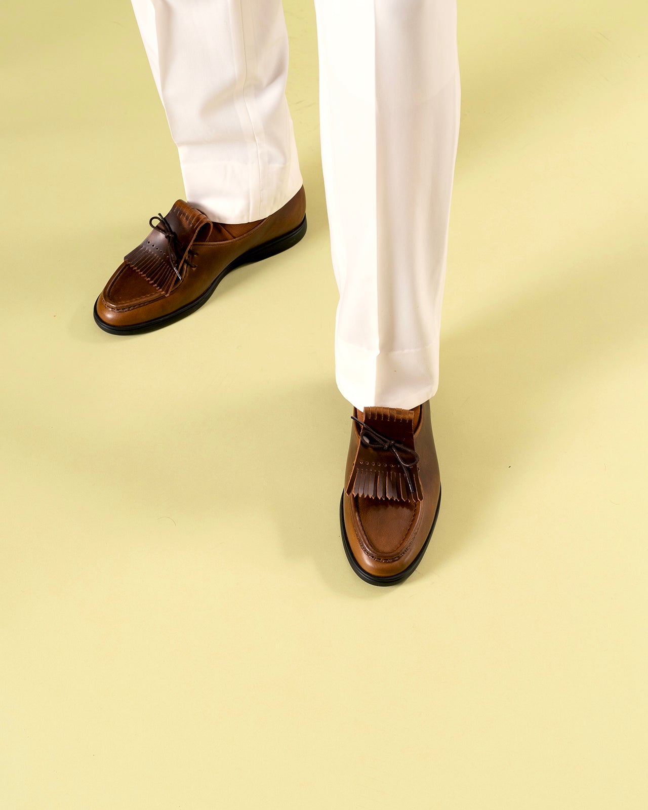 Relaxed brown loafers with tassels worn with white pants on a pistachio green background Timothée Paris