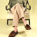 A man seated in a vintage iron chair wearing Timothée Paris handcrafted cognac leather apron-toe mocassin shoes, crafted in Portugal using traditional construction methods, paired with beige tailored trousers.