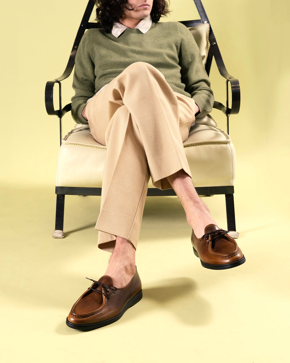 A man seated in a vintage iron chair wearing Timothée Paris handcrafted cognac leather apron-toe mocassin shoes, crafted in Portugal using traditional construction methods, paired with beige tailored trousers.