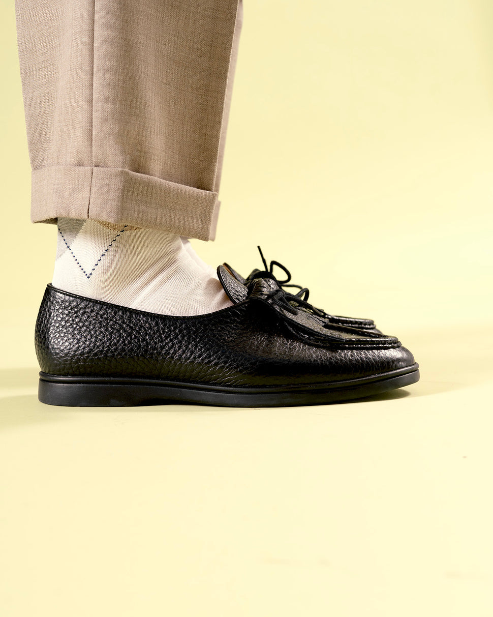 Person wearing black Beaune loafer mocassin shoes with rubber sole and white argyle socks on a light yellow background side view
