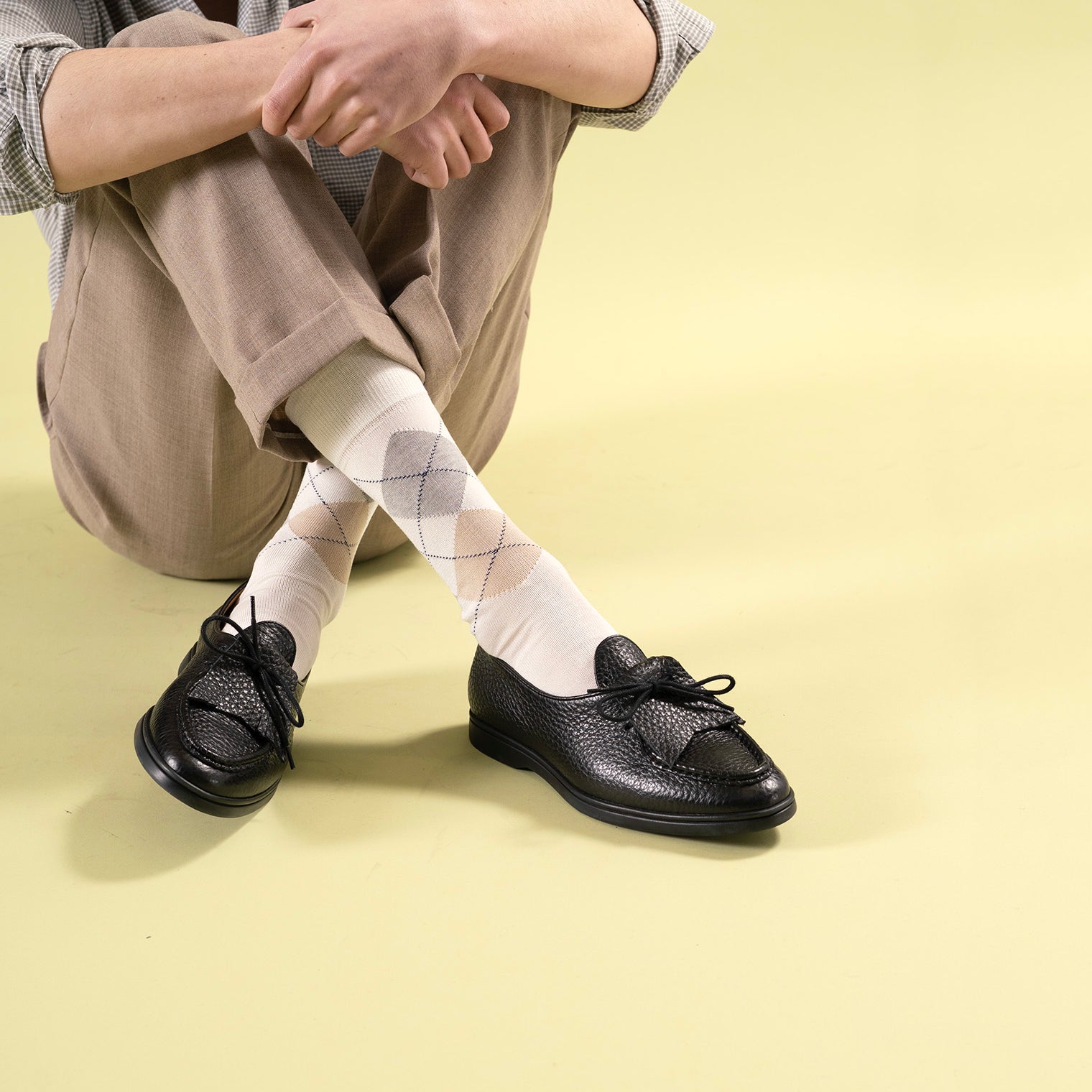 Person wearing black Beaune loafer mocassin shoes with rubber sole and white argyle socks on a light yellow background