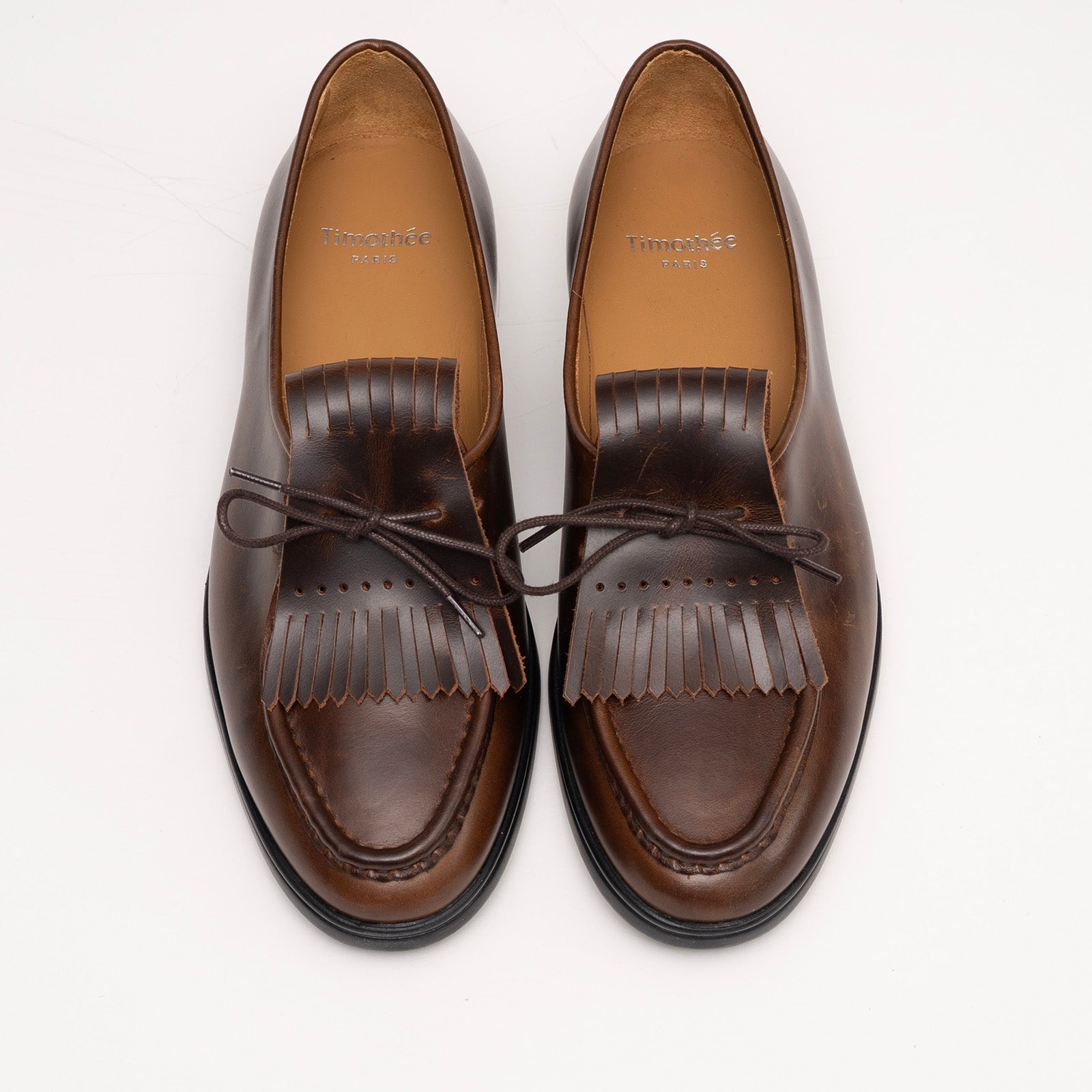 Pair of cognac brown loafers with fringes by Timothée Paris on a white background top down view 