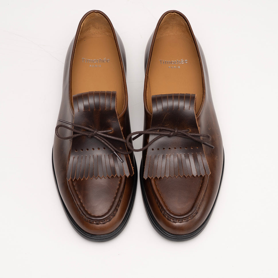 Pair of cognac brown loafers with fringes by Timothée Paris on a white background top down view 