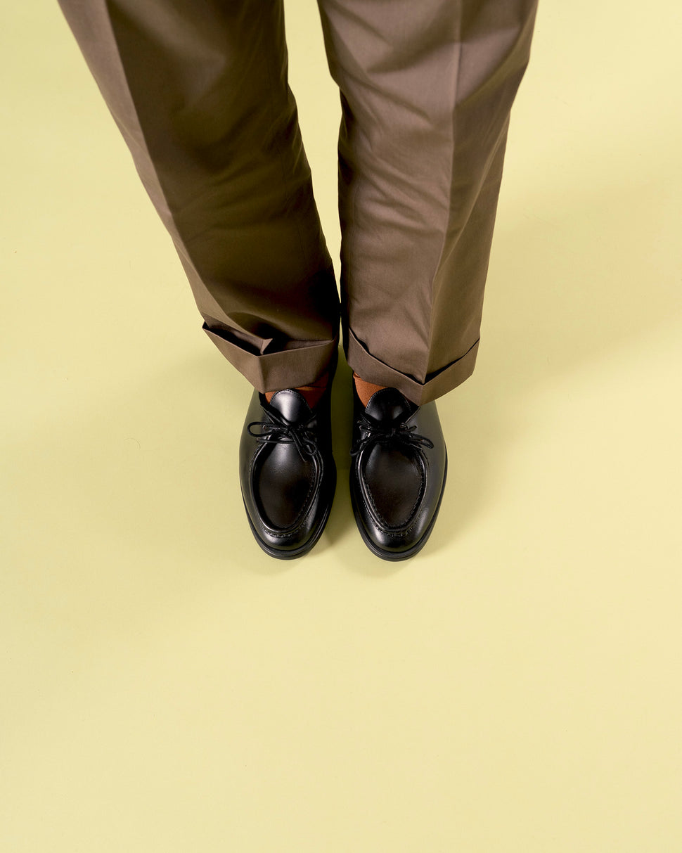 Top-down view of Timothée Paris black leather apron-toe mocassin shoes Beaune with fine lace detail, paired with cuffed dark -brown trousers and rust socks on a yellow-green surface