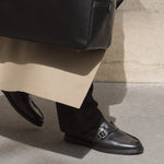 A close-up of a person wearing black leather penny loafers with a silver buckle, black trousers, and a beige coat, standing on a concrete surface next to a black leather bag.