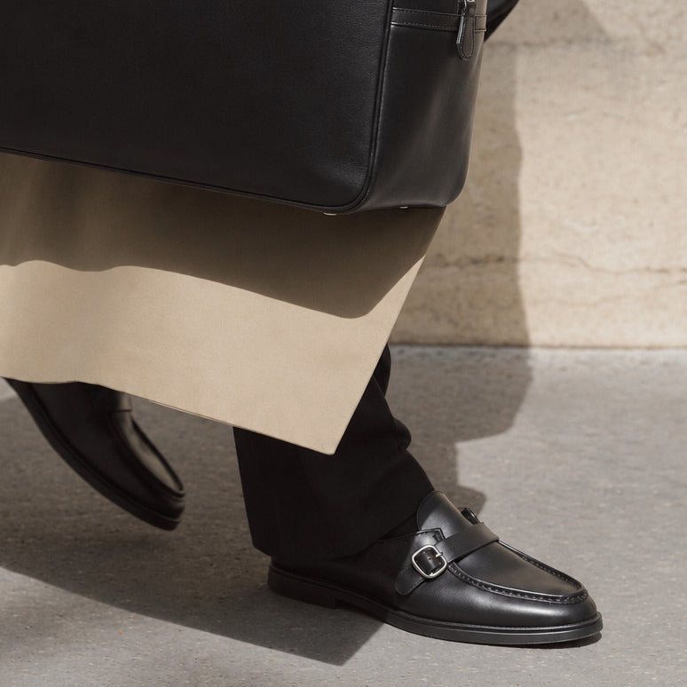 A close-up of a person wearing black leather penny loafers with a silver buckle, black trousers, and a beige coat, standing on a concrete surface next to a black leather bag.