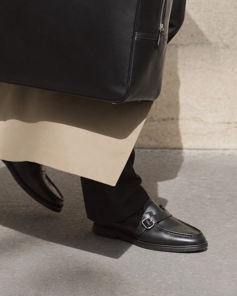 A close-up of a person wearing black leather penny loafers with a silver buckle, black trousers, and a beige coat, standing on a concrete surface next to a black leather bag.