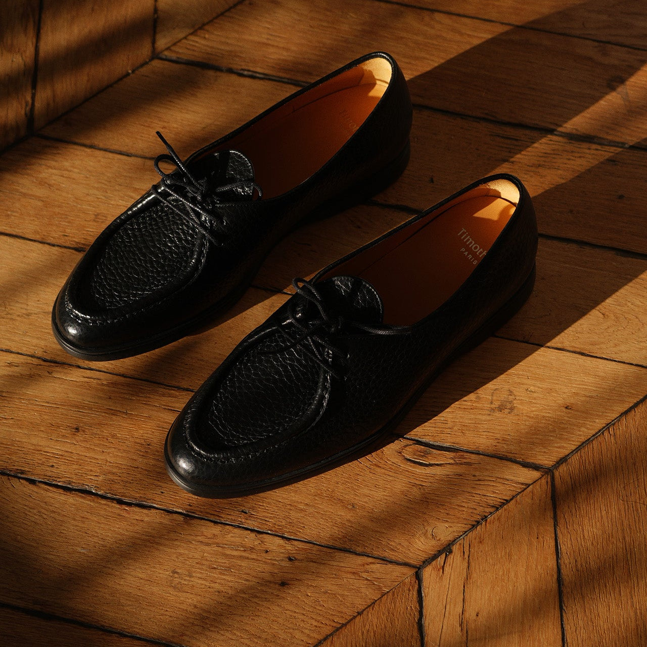Pair of black loafers by Timothée Paris on a wooden floor with warm lighting