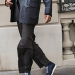A person walking outdoors wearing a dark navy coat, dark patterned trousers, and blue sneakers with white laces and white soles, set against a beige stone building wall.