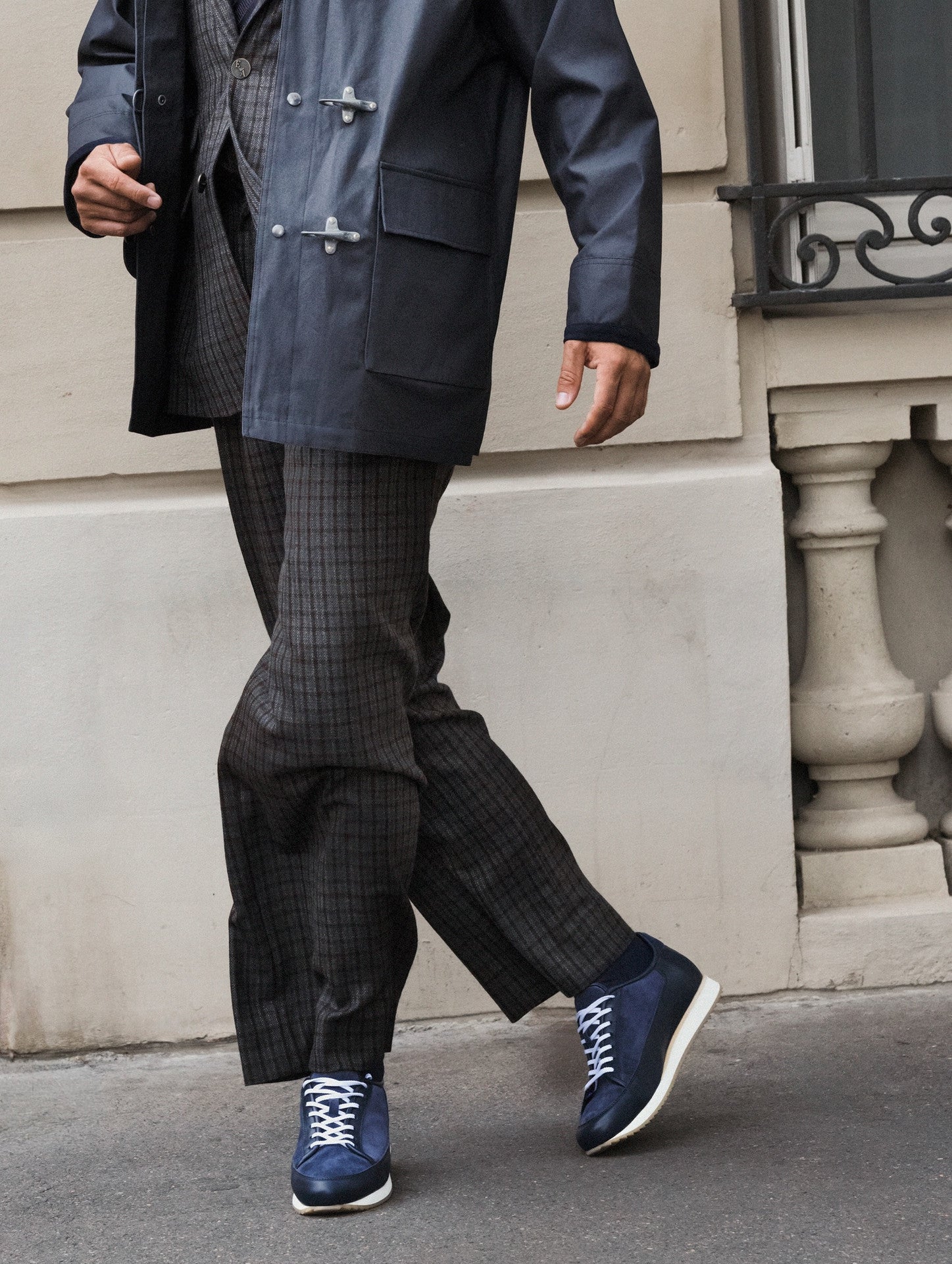 A person walking outdoors wearing a dark navy coat, dark patterned trousers, and blue sneakers with white laces and white soles, set against a beige stone building wall.