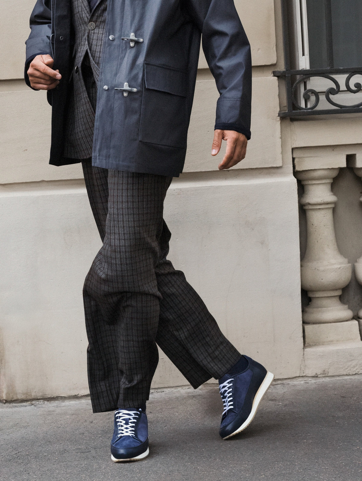 A person walking outdoors wearing a dark navy coat, dark patterned trousers, and blue sneakers with white laces and white soles, set against a beige stone building wall.